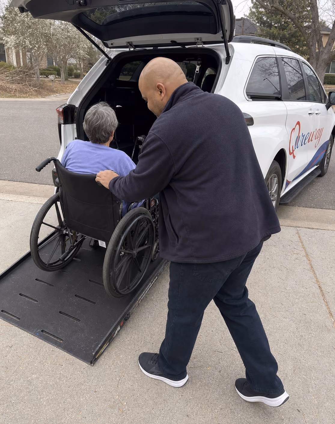 A Qureway driver assisting a wheelchair rider toward the van's open rear ramp